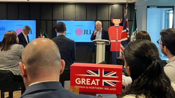 Un representante se dirige a un selecto grupo de asistentes en un auditorio exclusivo, respaldado por pantallas que exhiben la marca JLL y subrayan pilares clave: activos inmobiliarios de alto valor y sustentabilidad. El entorno está cuidadosamente ambientado con emblemas distintivos del Reino Unido, como la bandera británica y figuras icónicas, proyectando solidez internacional. En primer plano, una elegante caja con el logotipo GREAT BRITAIN & NORTHERN IRELAND refuerza el enfoque en oportunidades premium para inversores y compradores que buscan participar en los mercados inmobiliarios más dinámicos, sostenibles y prestigiosos a nivel global.