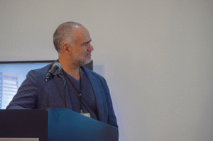 Un líder experimentado del sector inmobiliario premium, de cabello canoso y textura impecable, se posiciona con firmeza tras el atril principal. Elegante en un blazer oscuro y con credencial institucional al cuello, proyecta seguridad mientras se dispone a abordar las últimas tendencias en innovación arquitectónica y proyectos exclusivos de alto valor. Su presencia anticipa una visión vanguardista y estratégica dirigida a inversores y compradores que buscan excelencia, exclusividad e innovación en desarrollos inmobiliarios de primer nivel. El entorno sobrio enfoca toda la atención en su experiencia y liderazgo.