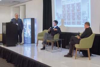 Tres destacados expertos del sector participaron en un panel sobre Innovación en Arquitectura, enfocado en el desarrollo de espacios urbanos excepcionales. Uno de los ponentes lideró la presentación desde el podio, mientras que los otros dos compartieron sus perspectivas desde el escenario, cada uno con micrófono. Una proyección de un edificio icónico ilustró la discusión, resaltando las tendencias actuales y las oportunidades exclusivas que ofrece la arquitectura de vanguardia para inversores y compradores inmobiliarios premium que buscan activos distintivos en entornos urbanos privilegiados.