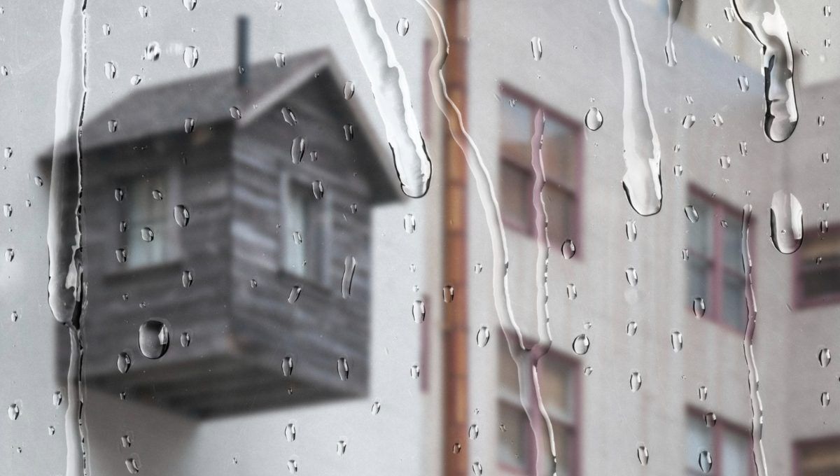 Vista a través de un ventanal donde las gotas de lluvia se deslizan sutilmente, ofreciendo una atmósfera serena y exclusiva. Al fondo, se distingue -aunque suavemente desenfocada- una terraza de madera integrada a una vivienda de tonos claros, reflejo del diseño contemporáneo y elegante propio del sector premium. La escena diurna bajo la llovizna resalta la privacidad y el confort del espacio, atributos ideales para quienes buscan invertir en propiedades excepcionales que combinan sofisticación arquitectónica con ambientes acogedores incluso en días lluviosos.