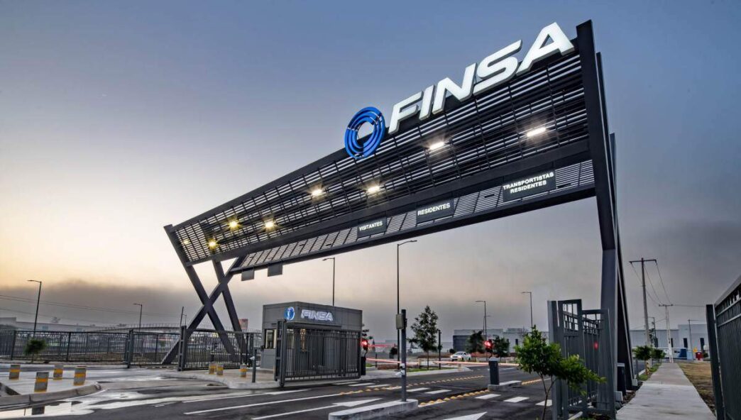 Acceso principal a parque industrial de alta gama, con un imponente letrero negro FINSA iluminado, caseta de seguridad equipada, barreras automáticas y vialidades completamente pavimentadas. Esta infraestructura moderna refleja los más altos estándares inmobiliarios para 2024 y subraya el compromiso con la Certificación LEED, garantizando eficiencia, sostenibilidad y máxima seguridad para inversionistas y compradores que exigen excelencia.