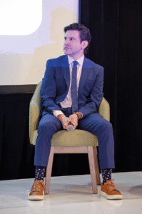 Un hombre elegantemente vestido con un traje azul, calcetas de diseño y sneakers marrones ocupa un asiento verde mientras sostiene un micrófono. Su atención se dirige hacia su izquierda, en una escena cuidadosamente ambientada con fondo de pantalla y cortina negra. El escenario es ideal para presentar oportunidades exclusivas de inversión en el sector hotelero premium de Puebla, permitiendo a potenciales compradores e inversionistas analizar propuestas inmobiliarias que destacan por su sofisticación, alta rentabilidad y ubicación estratégica en uno de los destinos más atractivos del país.