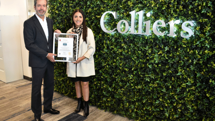 Celebración 30 años Colliers México: Dos personas sostienen un certificado frente al logo de Colliers sobre un fondo de follaje verde.