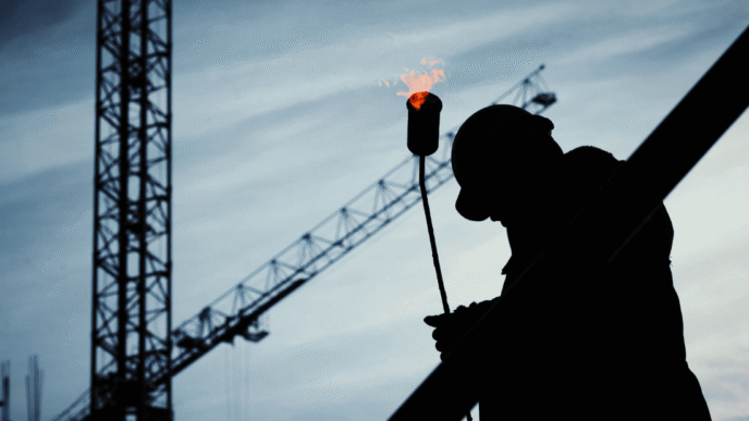 Silueta de obrero con soplete en construcción industrial de Aguascalientes. Crecimiento en actividad industrial visible con grúas de fondo. 125 chr