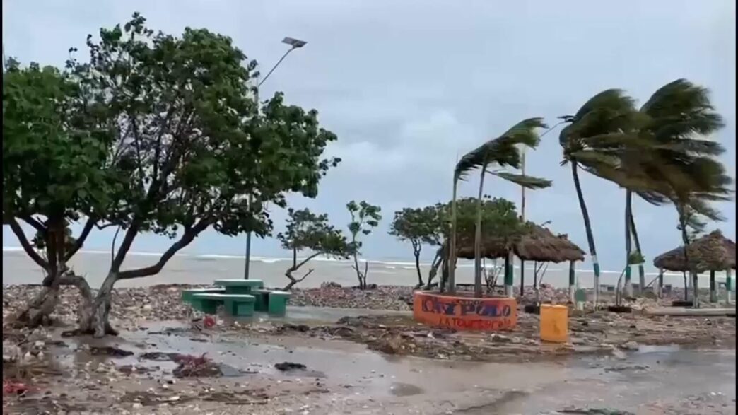 city/destination
Playa azotada por el viento en [Ciudad]. Árboles y palapas se inclinan por la fuerza del viento. Clima adverso en la [Ciudad].