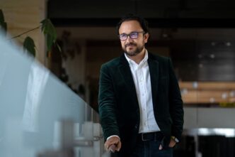 Hombre con saco verde, BBVA México. Predicción de 39 mil hipotecas. Rostro sonriente, lentes. Iluminación natural, actitud reflexiva y profesional.