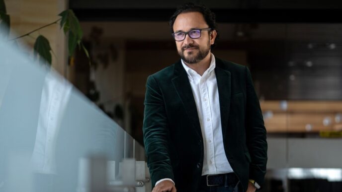 Hombre con saco verde, BBVA México. Predicción de 39 mil hipotecas. Rostro sonriente, lentes. Iluminación natural, actitud reflexiva y profesional.