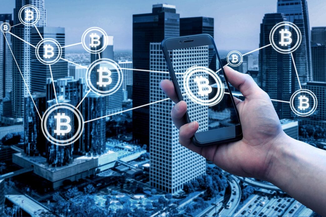 Mano sujetando un teléfono mostrando Bitcoin sobre un fondo urbano, representando la revolución del blockchain en el real estate.