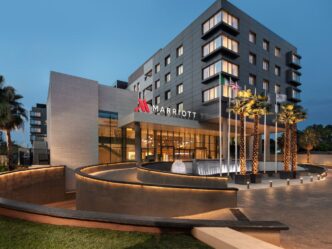 Fachada del Marriott, una cadena hotelera líder mundial, con palmeras y banderas ondeando. Iluminación nocturna destaca arquitectura moderna y elegante.