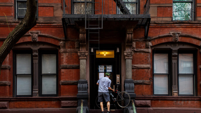 Fachada de edificio de ladrillo en Nueva York, con un hombre subiendo las escaleras con una bicicleta. Impacto inmobiliario y leyes de vivienda.