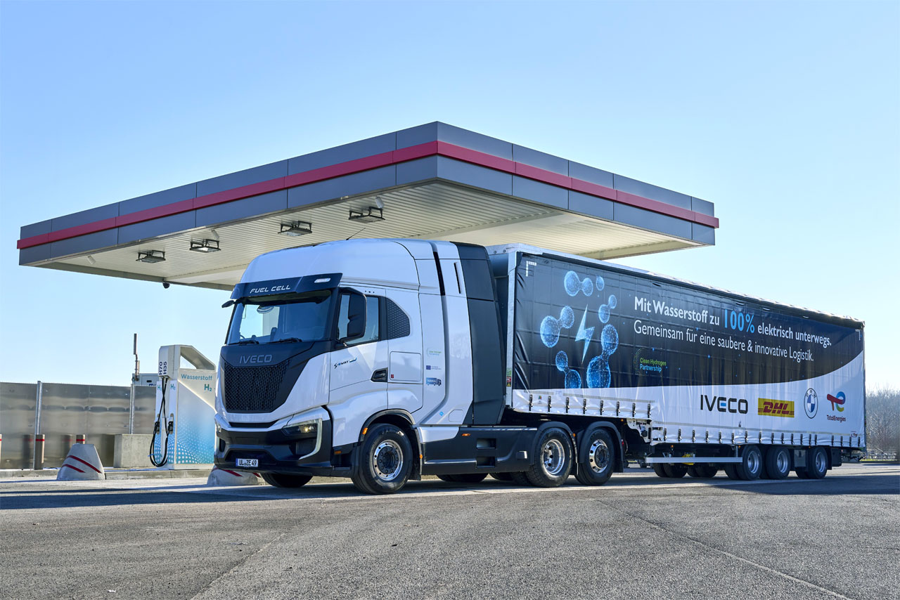 Camión de hidrógeno IVECO en estación de servicio, promoviendo el futuro del transporte sostenible. Diseño innovador y logotipos de colaboración.