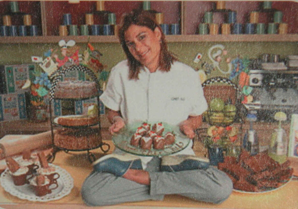 speakers/headshot
Chef sonriendo, sentada con las piernas cruzadas, sosteniendo un plato de postres. Variedad de pasteles y dulces alrededor.