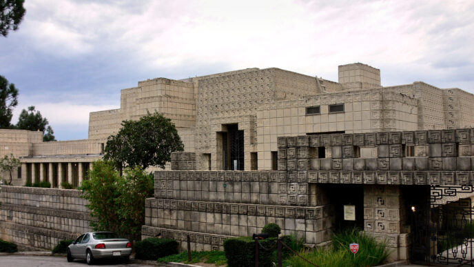 Vista exterior Casa Ennis, LA. Arquitectura maya centenaria con bloques texturizados. Auto estacionado y vegetación circundante. Diseño icónico de Frank Lloyd Wright.