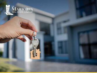 Llaves de nueva oferta inmobiliaria 2022. Mano sosteniendo llave con llavero de casa frente a una vivienda moderna. Colocación inmobiliaria.