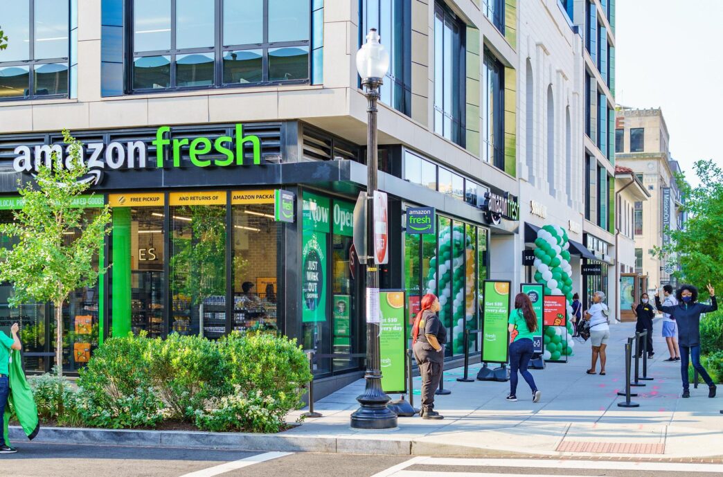 Tienda Amazon Fresh en Glendale con gente esperando. Estrategias de mercado en desinversión y oportunidades visibles. Fachada moderna y letreros verdes.