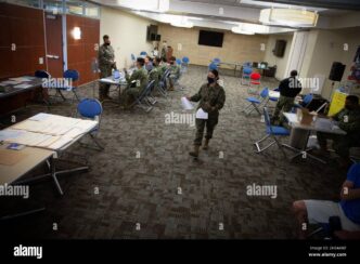 Militares procesando documentos en un salón amplio, usando mascarillas. Escritorios y sillas azules. Suelo con patrón geométrico. Iluminación brillante.