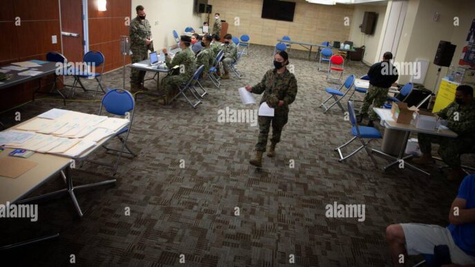 Militares procesando documentos en un salón amplio, usando mascarillas. Escritorios y sillas azules. Suelo con patrón geométrico. Iluminación brillante.