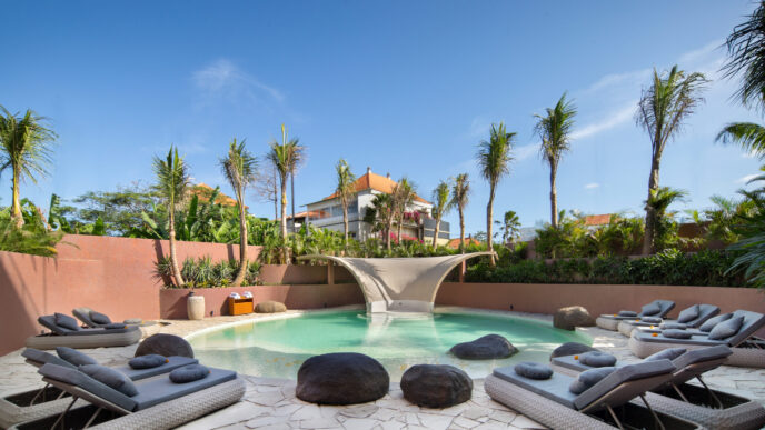 Piscina de hotel sostenible en Bali con tumbonas, rocas y palmeras. Un oasis de relajación comprometido con la sostenibilidad hotelera en Bali.