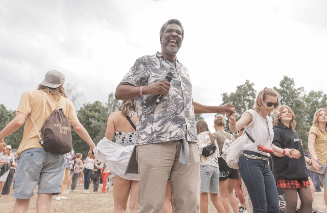 Orador interactúa con el público en conferencia. Evento al aire libre con música y baile. Gente tomándose de las manos.