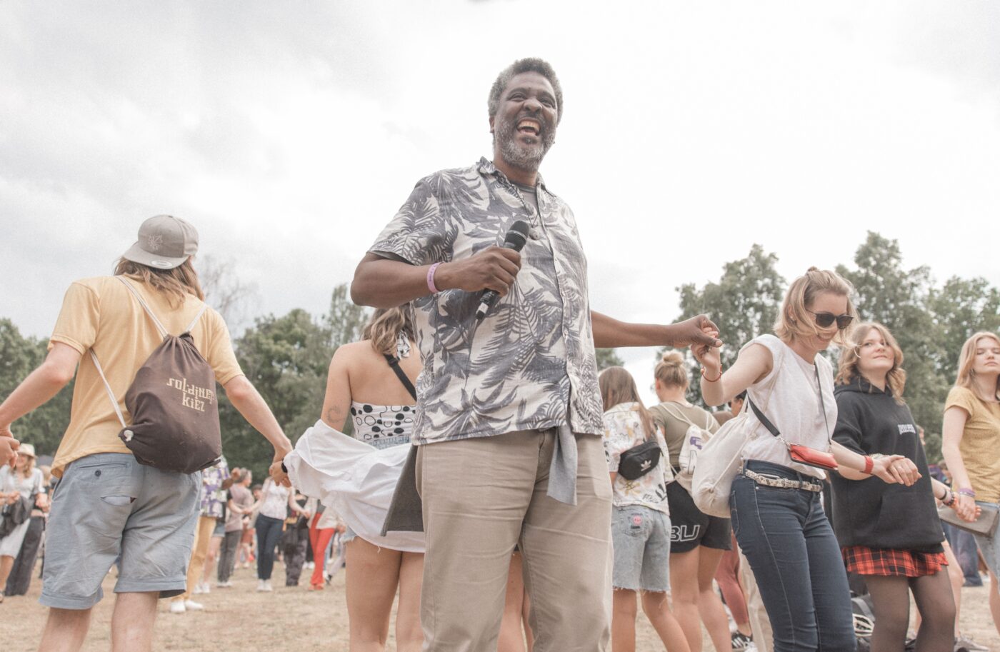 Orador interactúa con el público en conferencia. Evento al aire libre con música y baile. Gente tomándose de las manos.