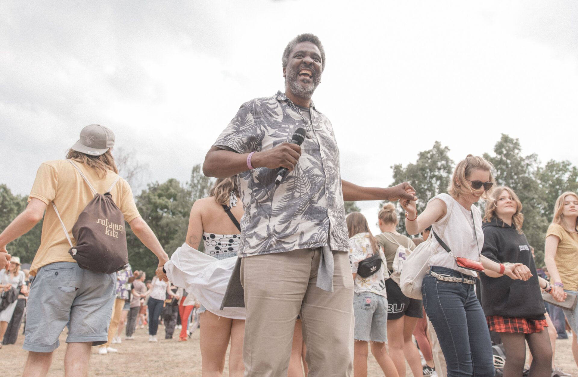 Orador interactúa con el público en conferencia. Evento al aire libre con música y baile. Gente tomándose de las manos.