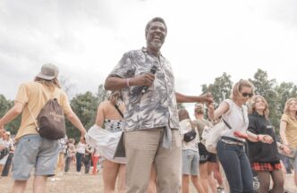 Orador interactúa con el público en conferencia. Evento al aire libre con música y baile. Gente tomándose de las manos.