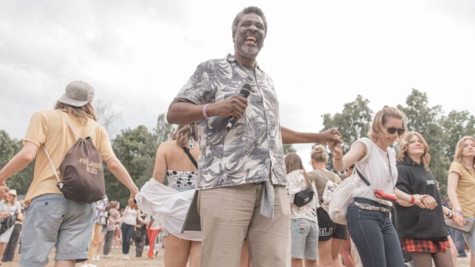 Orador interactúa con el público en conferencia. Evento al aire libre con música y baile. Gente tomándose de las manos.