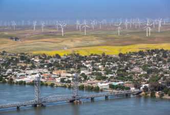 Vista aérea de un parque eólico offshore con molinos generando energía eólica. Impacto en el real estate visible con la ciudad y el puente.