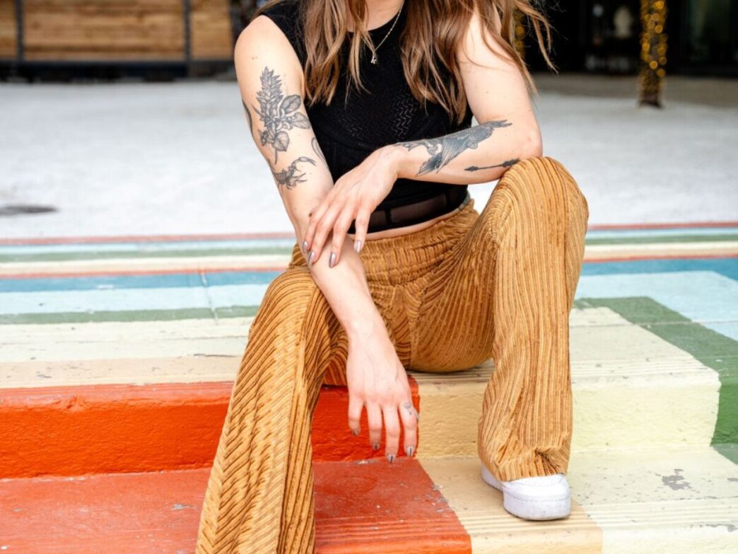 Mujer sentada en escaleras coloridas con pantalones acampanados y tatuajes en brazos. Estilo moderno en Chicago, como en Bar Bambi. 124 carácteres