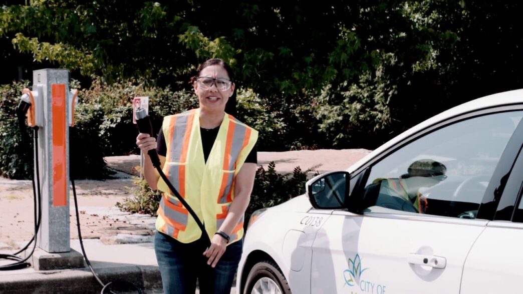 Mujer con chaleco reflectante sosteniendo conector para carga de vehículo eléctrico en Vancouver. Impulso sostenible para electrificación de flotas.