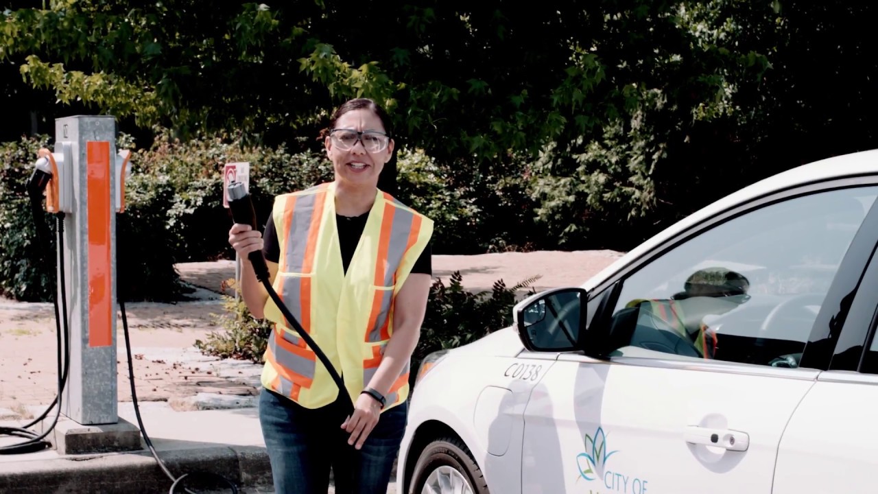 Mujer con chaleco reflectante sosteniendo conector para carga de vehículo eléctrico en Vancouver. Impulso sostenible para electrificación de flotas.