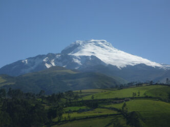 Vista panorámica del nevado Chimborazo, con su cumbre cubierta de nieve brillante, resaltando sobre las colinas verdes y el cielo azul despejado.