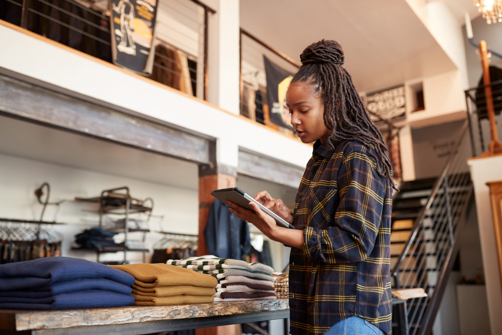 Here are some possible alt text options:

**Option 1 (Focus on the woman and her activity):**

Mujer usando tablet en tienda de ropa. Gestión de inventario y ventas. Tecnología en el comercio minorista.

**Option 2 (Focus on the store environment):**

Tienda de ropa moderna con mujer trabajando en tablet. Ambiente de compras y gestión de stock.

**Option 3 (More general description):**

Mujer revisando inventario en una tienda de ropa con una tablet. Gestión moderna de negocios.