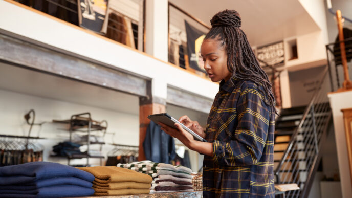 Here are some possible alt text options:

**Option 1 (Focus on the woman and her activity):**

Mujer usando tablet en tienda de ropa. Gestión de inventario y ventas. Tecnología en el comercio minorista.

**Option 2 (Focus on the store environment):**

Tienda de ropa moderna con mujer trabajando en tablet. Ambiente de compras y gestión de stock.

**Option 3 (More general description):**

Mujer revisando inventario en una tienda de ropa con una tablet. Gestión moderna de negocios.