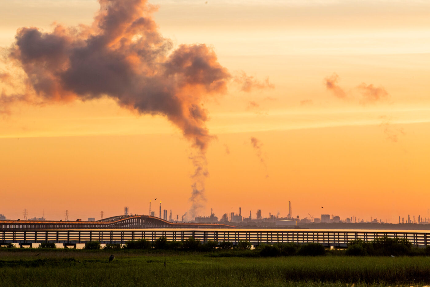Amanecer industrial en Texas: refinerías, puente y humo. Impacto LATAM visible en la expansión. Horizonte anaranjado contrasta con la actividad fabril.