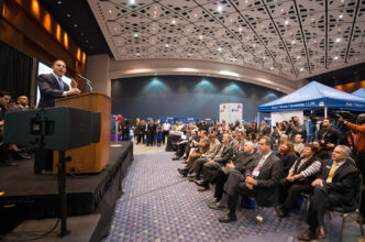 Un orador en un podio frente a una audiencia en una conferencia. El evento parece ser un evento corporativo grande y bien atendido.