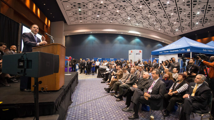 Un orador en un podio frente a una audiencia en una conferencia. El evento parece ser un evento corporativo grande y bien atendido.