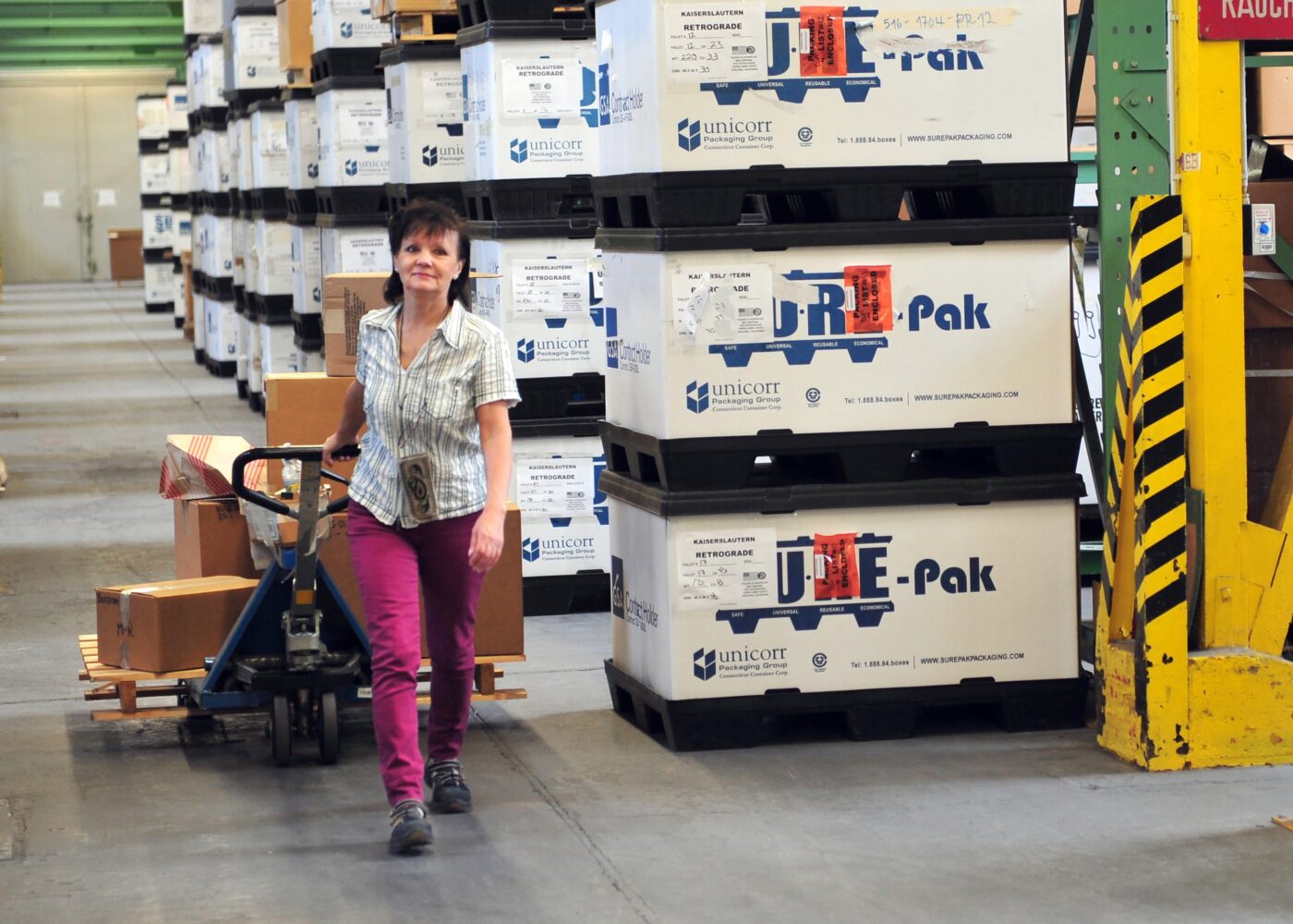 Mujer transportando cajas en almacén logístico con carretilla. Embalajes de Unicorr y JDE-Pak apilados. Operaciones de Fidelity en Europa.