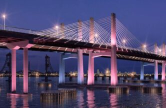Puente iluminado en New Jersey al anochecer. Pilares rosados reflejados en el agua. Impulso al sector industrial visible en la arquitectura urbana.