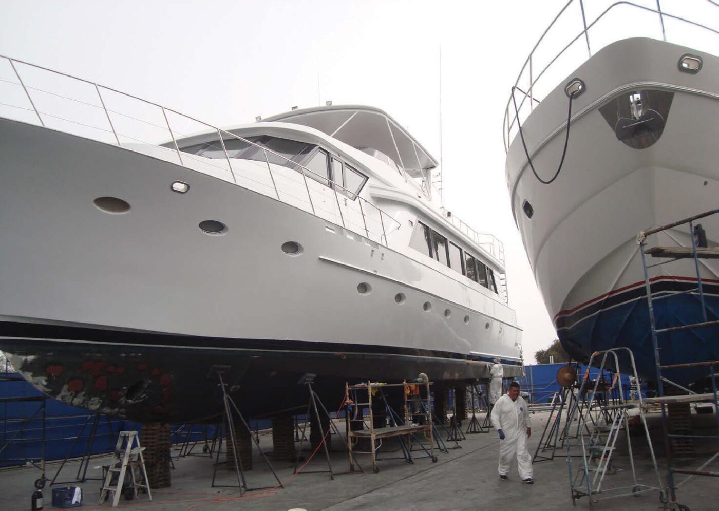 The image doesn't fit any of the specified categories. I will provide a description of the image instead.

Dos yates blancos en dique seco sobre soportes, con un hombre vestido de blanco caminando cerca.