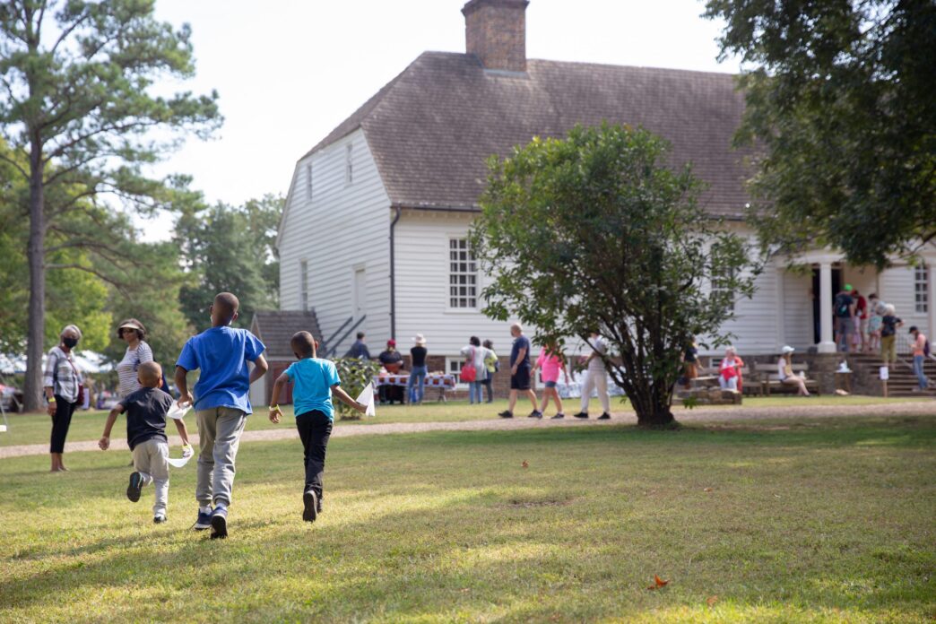 Tres niños corren en un campo verde hacia una casa blanca histórica. Fondos para la conservación del patrimonio, impacto en Real Estate.
