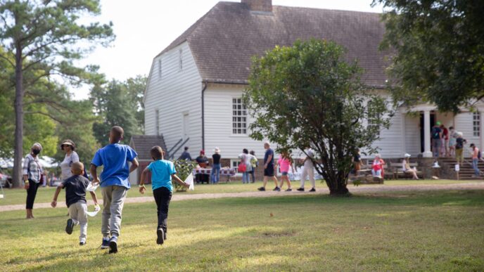 Tres niños corren en un campo verde hacia una casa blanca histórica. Fondos para la conservación del patrimonio, impacto en Real Estate.