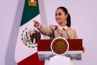 Claudia Sheinbaum en Oaxaca anuncia Fideicomisos para vivienda maestros. Mujer señalando, podio con escudo nacional y bandera de México.
