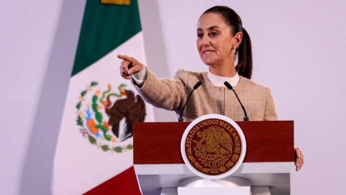 Claudia Sheinbaum en Oaxaca anuncia Fideicomisos para vivienda maestros. Mujer señalando, podio con escudo nacional y bandera de México.