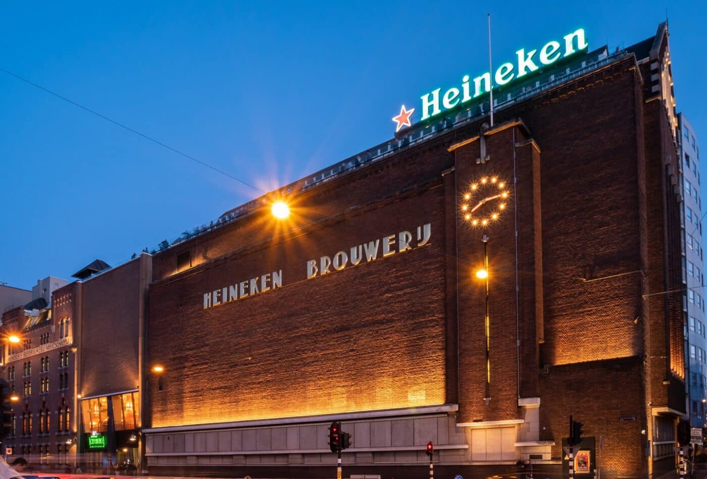 Fachada de la cervecería Heineken en Ámsterdam. Arquitectura de ladrillo con reloj iluminado y letrero de Heineken. Anuncia planta en Yucatán.