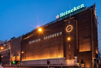Fachada de la cervecería Heineken en Ámsterdam. Arquitectura de ladrillo con reloj iluminado y letrero de Heineken. Anuncia planta en Yucatán.