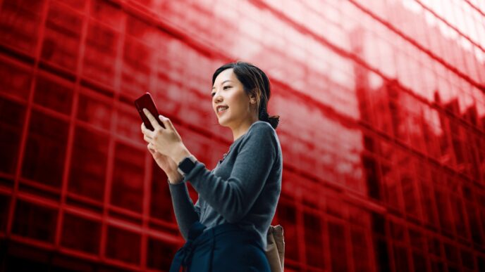 Mujer sonríe usando su celular frente a edificio rojo. Hipoteca HSBC Verde ofrece opciones de vivienda en México. Nuevo producto para tu hogar.