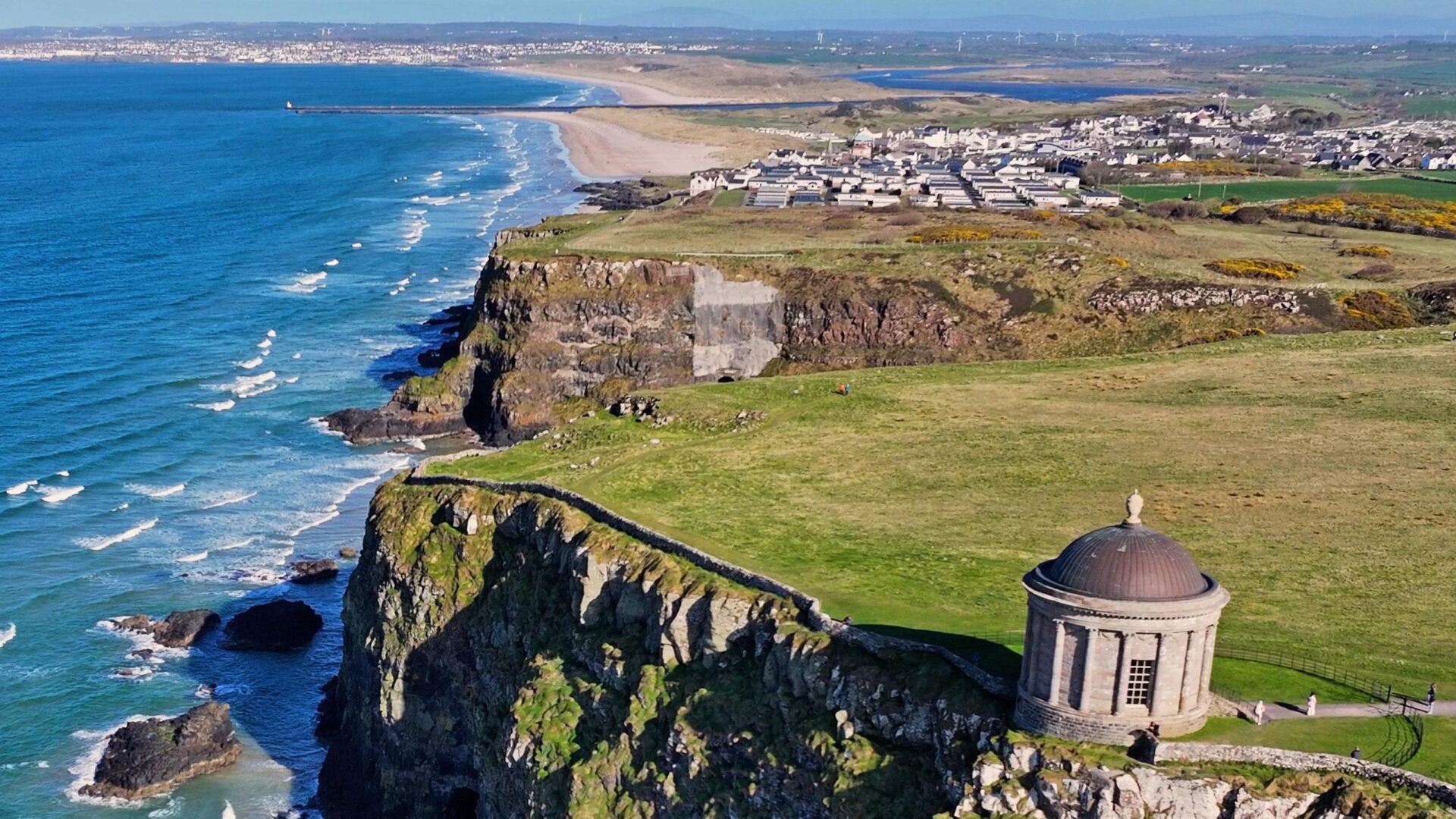 Vista aérea de la costa del Reino Unido con acantilados, playa y un templo, mostrando el impacto ambiental de la renovación costera. Detalle del paisaje.
