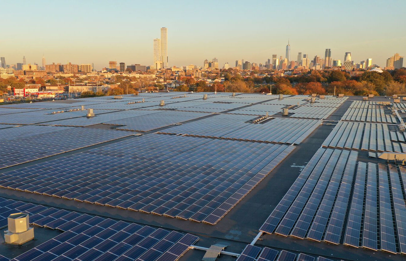 Paneles solares en techo de edificio logístico con horizonte urbano. Energía solar para real estate logístico, impacto y sostenibilidad. Vista aérea.