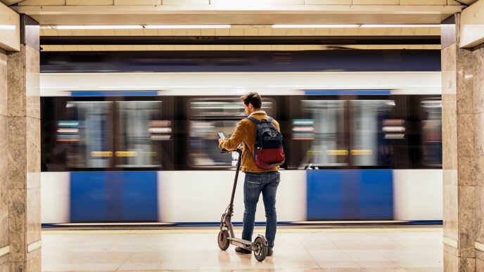 Hombre en patinete eléctrico en estación de tren, revisando su móvil con un tren borroso de fondo. Movilidad sostenible e inmobiliario ferroviario.